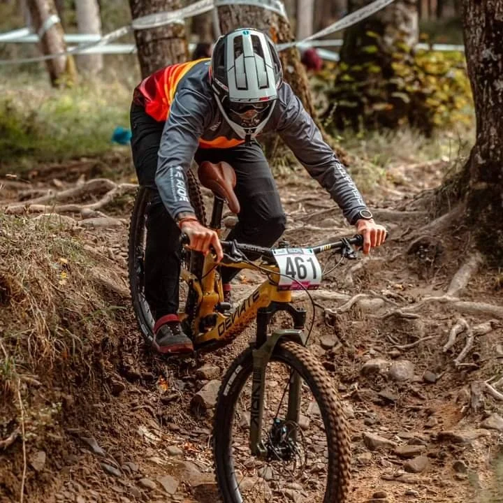 Mountain biker wearing helmet and custom race jersey navigating rough, root-filled trail in a forest.