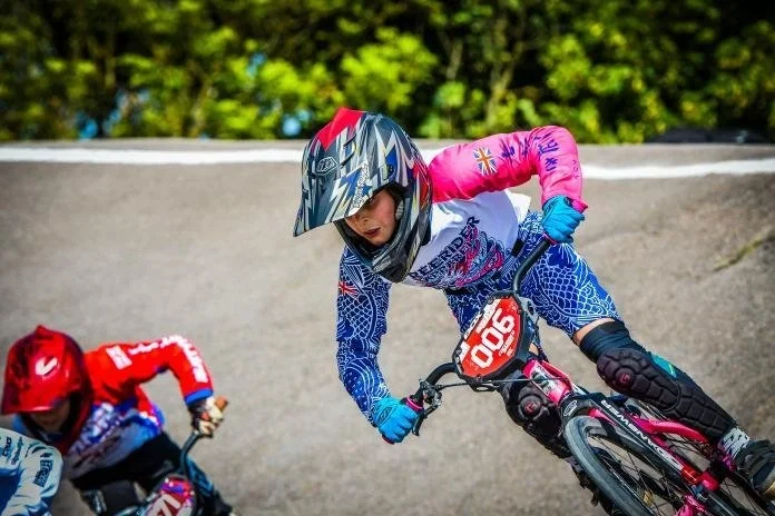 Two young girls racing on BMX bikes on a curved outdoor track, wearing helmets and colorful protective gear.