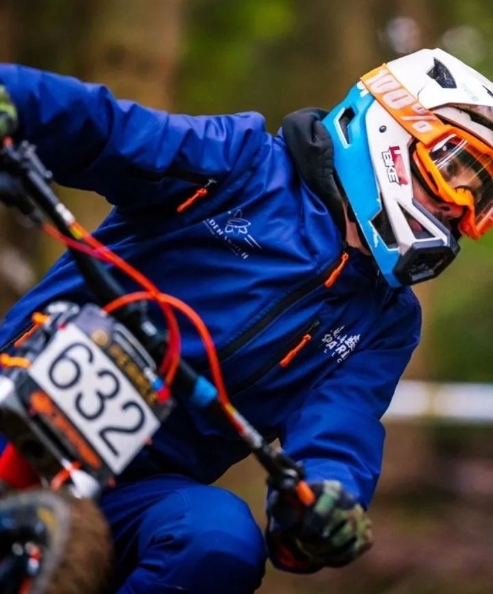 A person wearing blue protective gear and a full-face helmet riding a mountain bike wearing a waterproof MTB jacket on a trail.