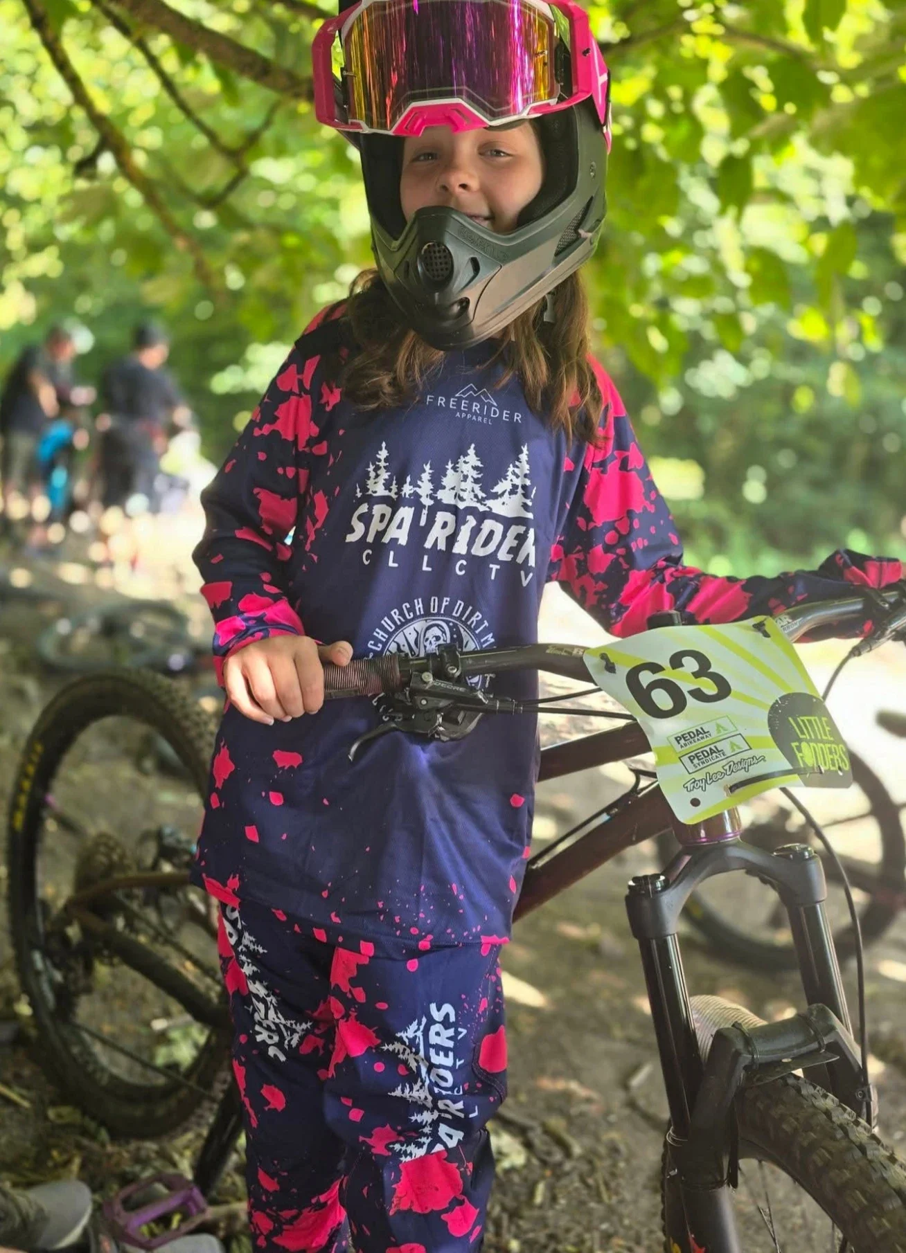 Girl wearing a helmet and custom down hill race kit inclusding jersey and pants by Freerider Apparel, standing with mountain bike, in a wooded area, with others in the background.