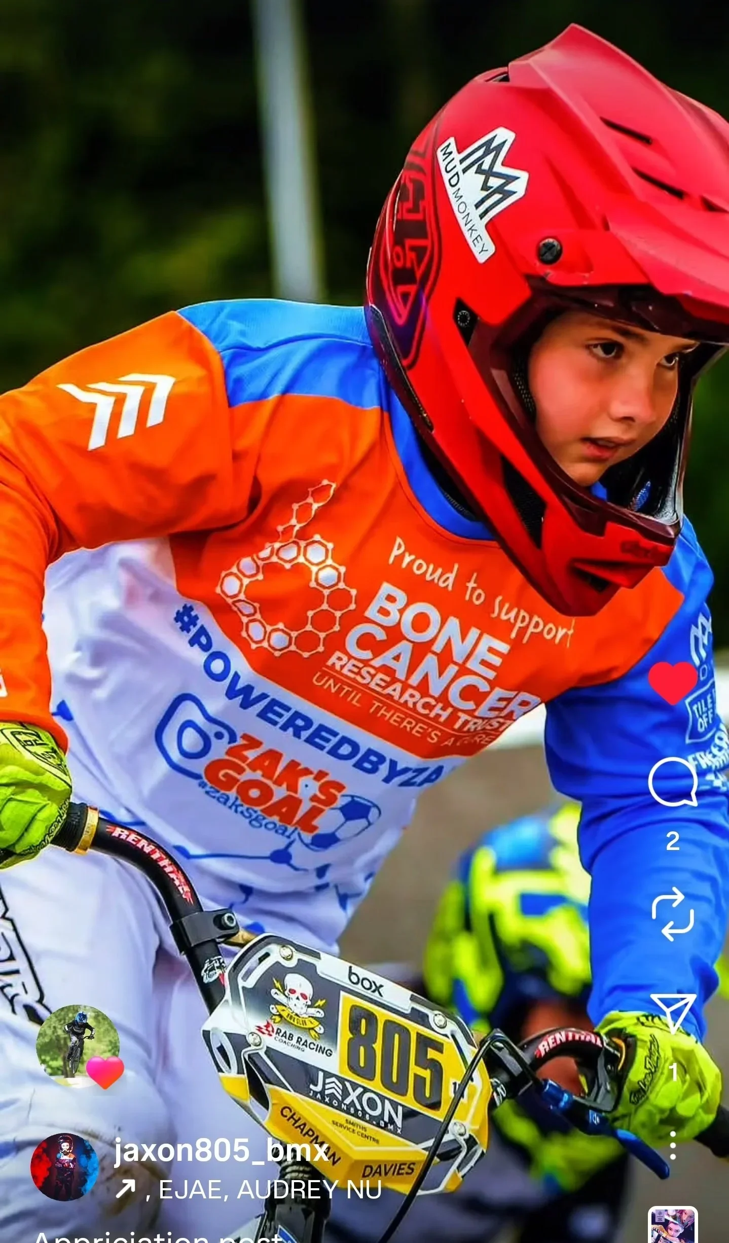 Young mountain biker with a red helmet and colorful custom designed jersey, participating in a race, with a noticeable number 805 on the bike, wearing yellow gloves, and a background of another rider in the distance.