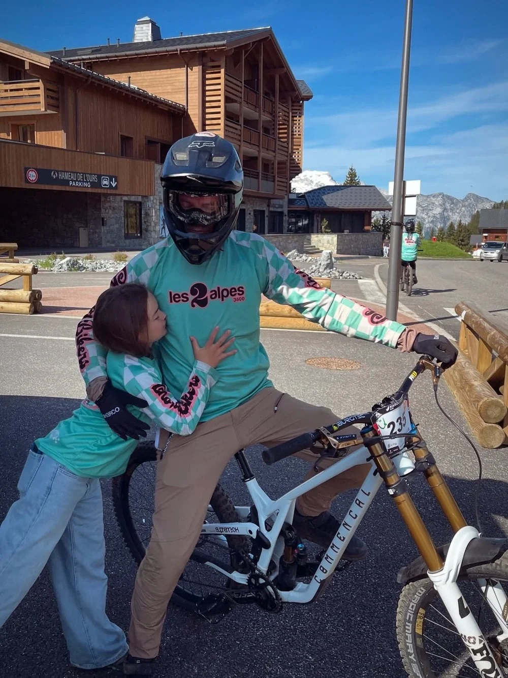 A person in a cycling helmet and riding gear on a mountain bike is being hugged by a young girl wearing a matching cycling jersey. They are outdoors near a wooden building with mountains in the background.