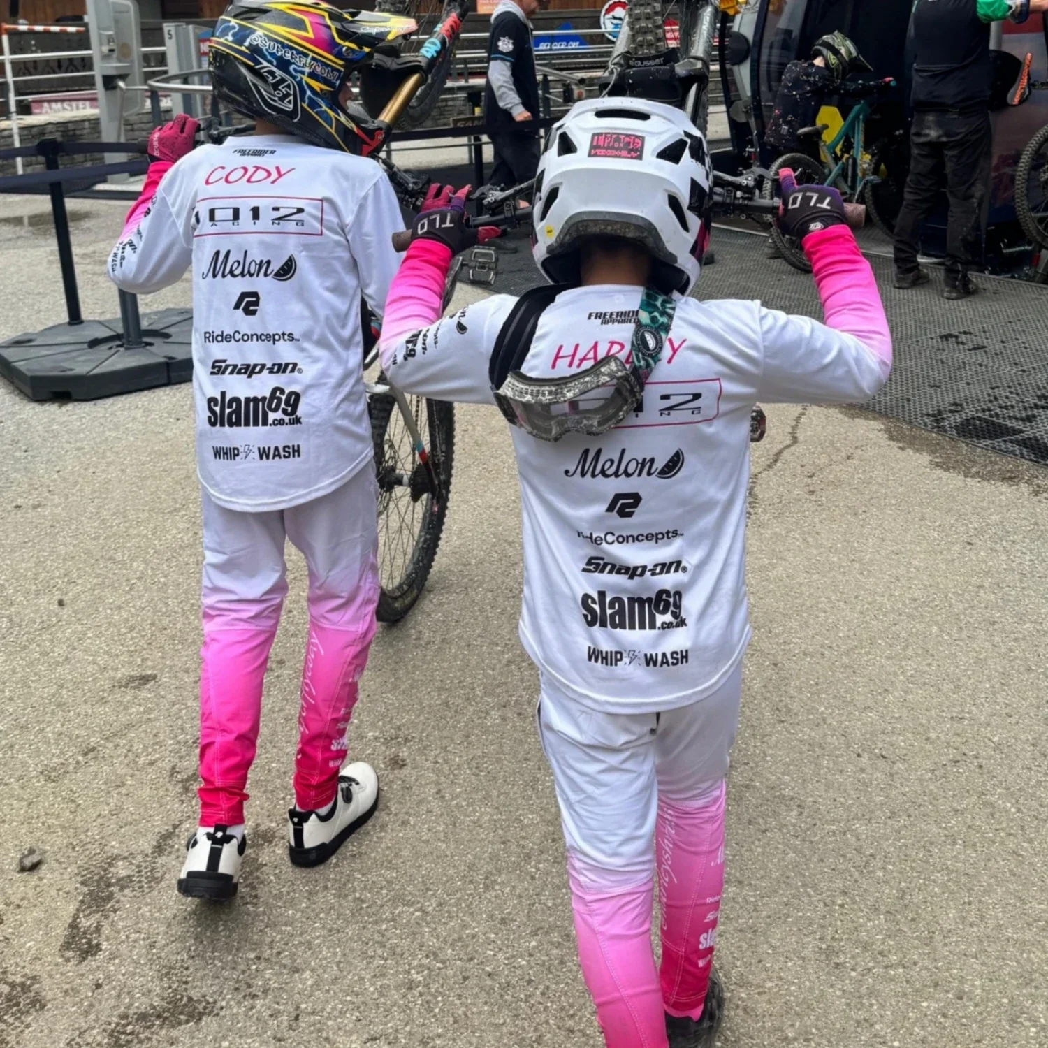 Two young riders in matching white and pink mountain bike gear standing with bikes in a pit area, preparing for a race.