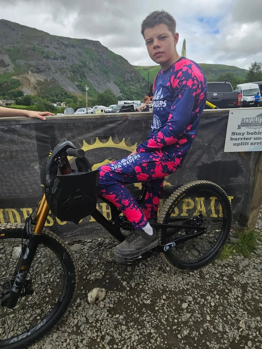 A young boy in a bright pink and blue Spa Riders Freerider Apparel outfit sitting on a black mountain bike, outdoors on gravel with a mountainous landscape in the background.