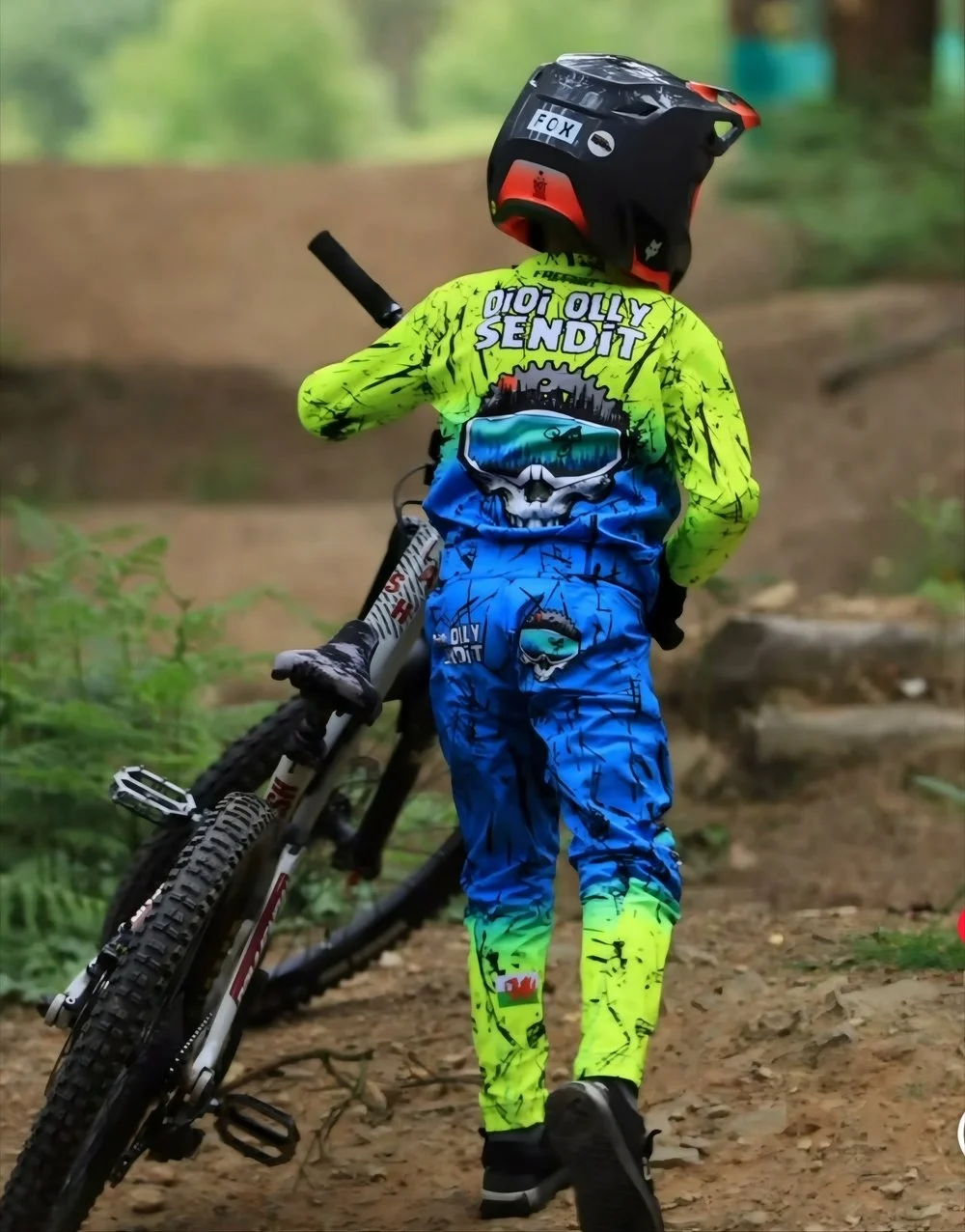 A young child wearing a mountain biking helmet and colorful  personolised custom Freerider Apparel gear is walking a mountain bike on a dirt trail in a wooded area.