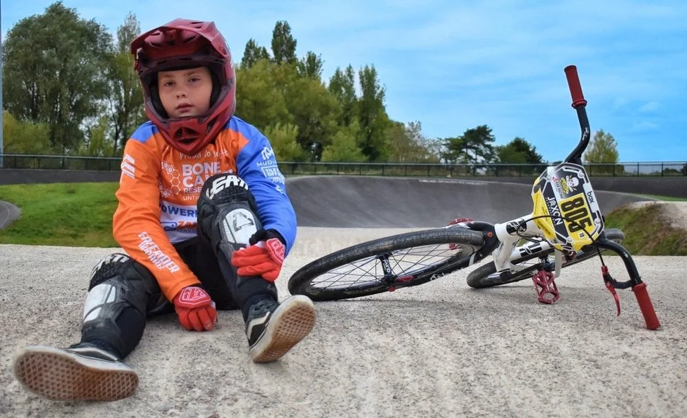 A young boy in a colorful custom designed bmx jersey jersey and helmet sitting on the ground next to a fallen BMX bike at a skate park on a cloudy day.