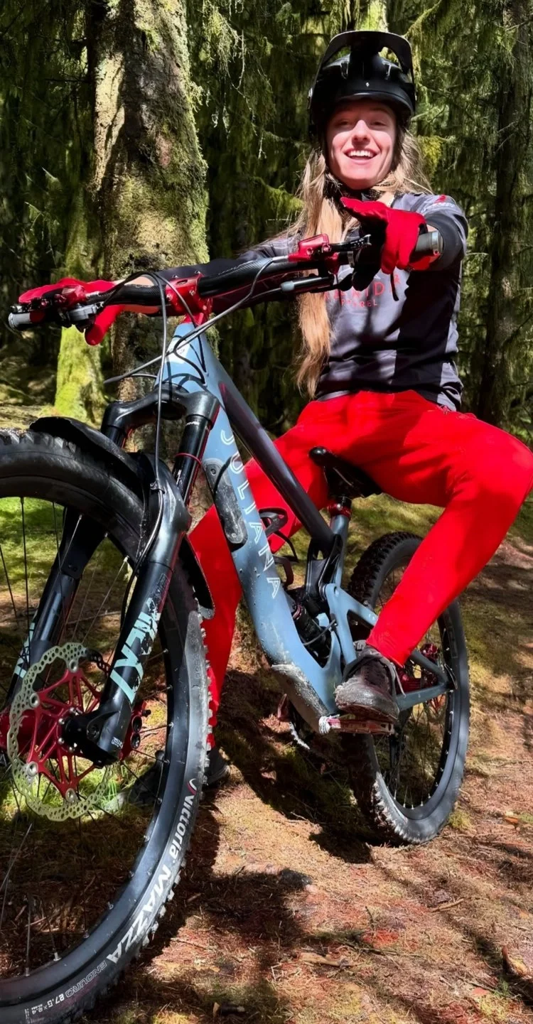 A smiling woman in outdoor gear, including a black helmet and red gloves, sitting on a mountain bike in a forest with green moss-covered trees.