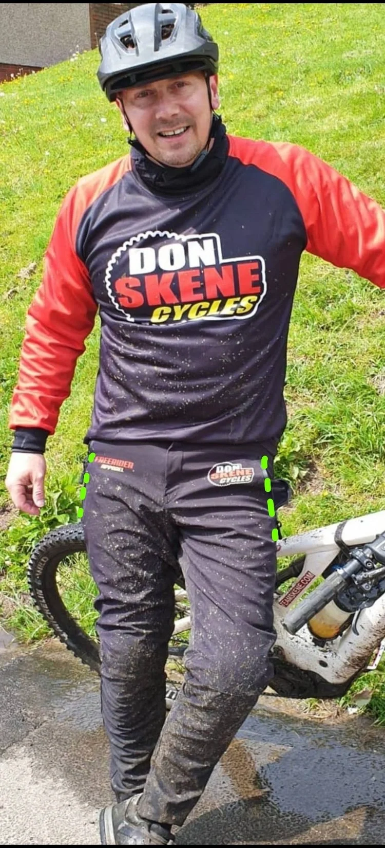 A man dressed in Don Skene Cycles cycling gear, wearing a protective helmet, standing next to a mountain bike, with mud and dirt on his clothing, outdoors on a grassy area.