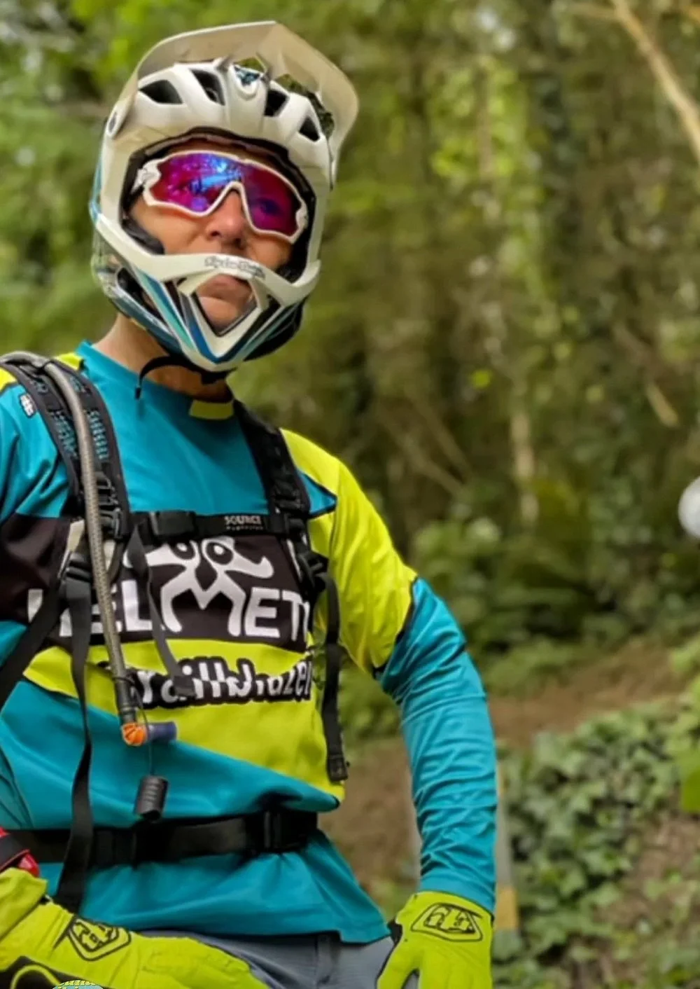 Mountain biker wearing a white helmet, purple-tinted goggles, blue and yellow long-sleeve jersey, yellow gloves, and a hydration pack stands in a green forested area.