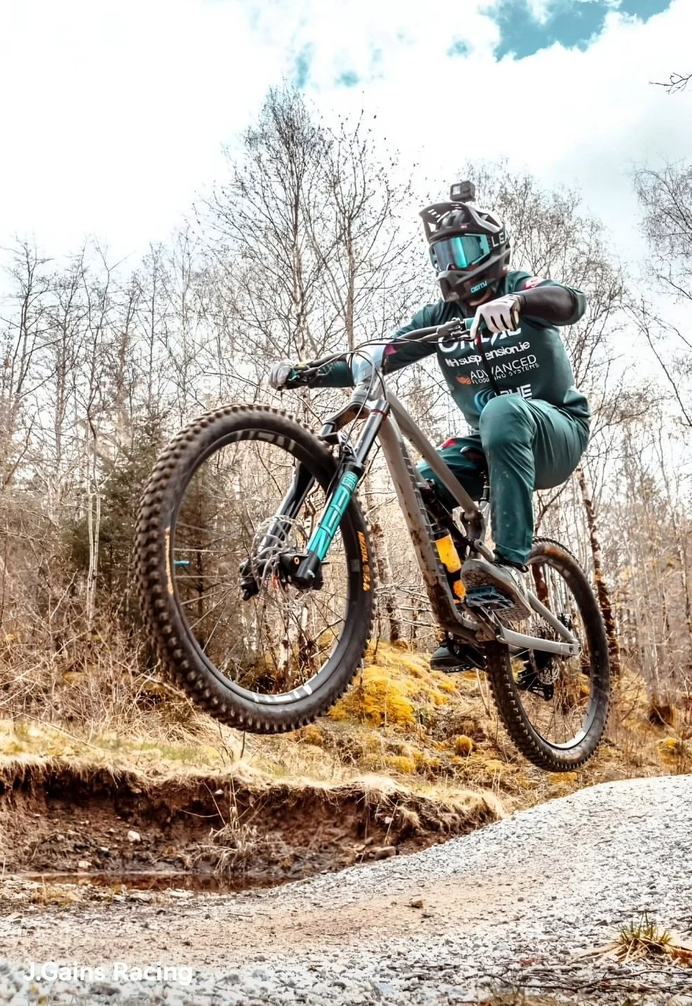 A mountain biker wearing a helmet and protective gear is airborne over a dirt trail in a forest during daytime.