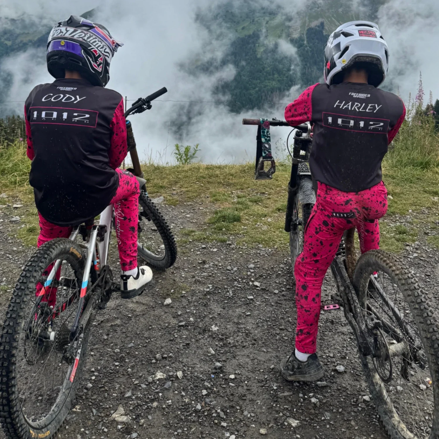Two young mountain bikers wearing black and pink gear with helmets, sitting on their bikes and looking at a smoky mountain landscape with fog and trees in the background.