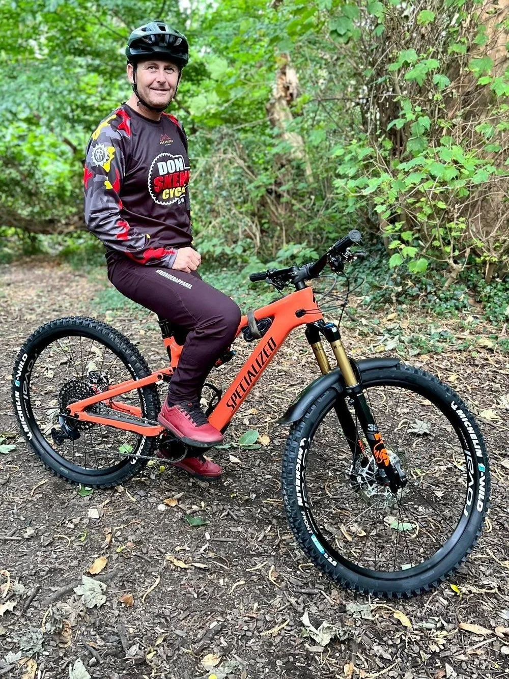 A man wearing a helmet and mountain biking gear sitting on an orange Specialized mountain bike on a dirt trail surrounded by green foliage.