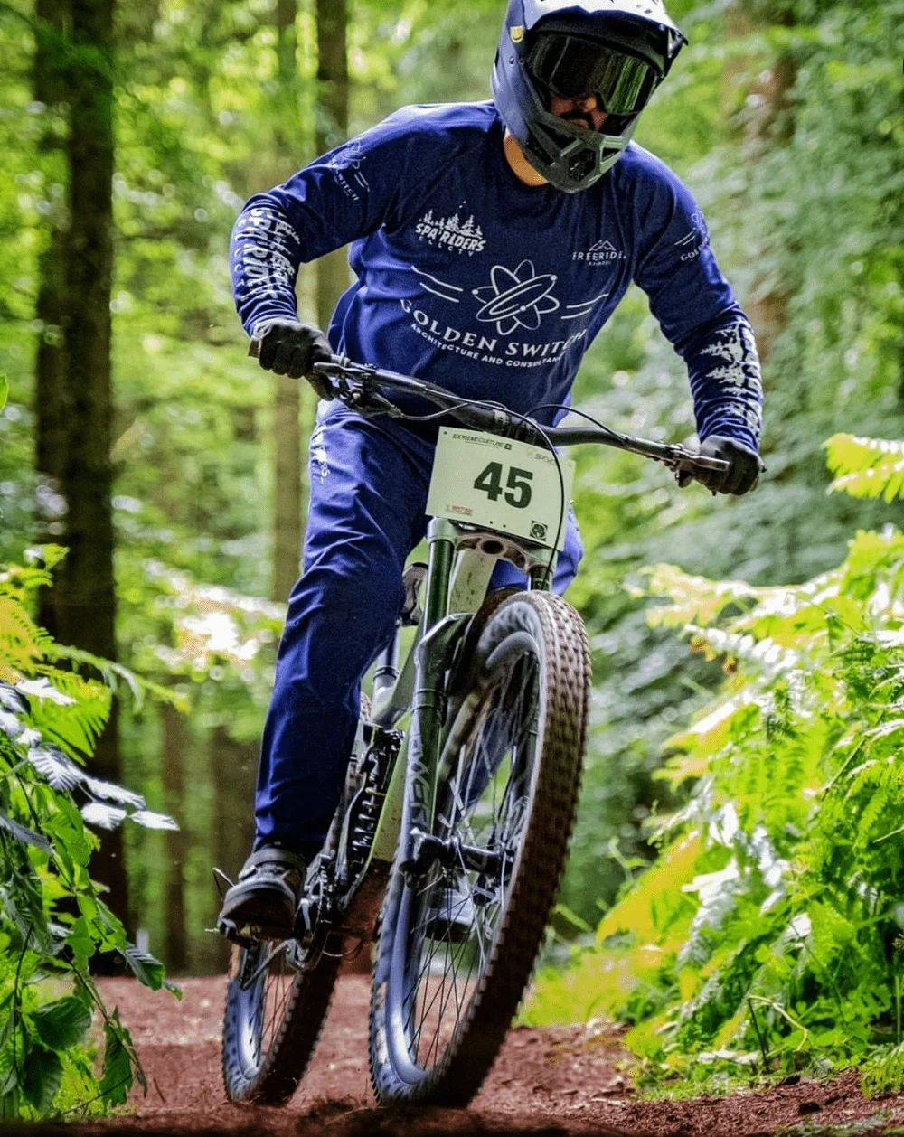 A mountain biker wearing a blue helmet, jacket, and pants riding a mountain bike on a dirt trail in a forest.