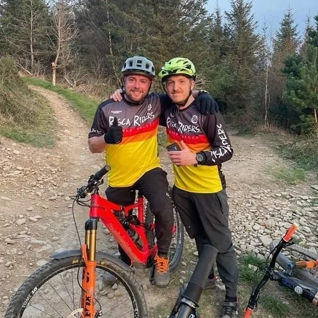 Two men in matching yellow and black Risca Riders bike jerseys and helmets, one sitting on a red mountain bike and the other standing beside him with his arm around him, on a dirt trail in a wooded area.