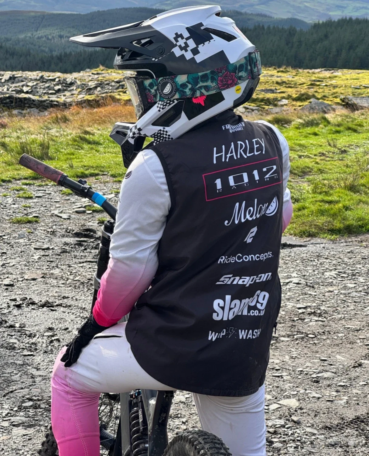 A person wearing a full-face helmet with various colorful designs on it, celebrating a mountain biking event, standing in an outdoor setting with green hills and trees in the background. The person is dressed in a black and white jersey with multiple