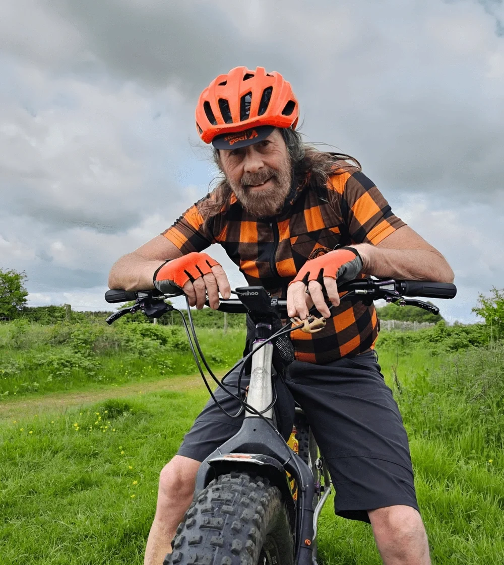 A man with gray hair and beard smiling while riding a black fat-tire mountain bike outdoors. He is wearing an orange helmet, orange gloves, a black and orange checkered cycling jersey, and black shorts. The background features green grass, shrubs, an