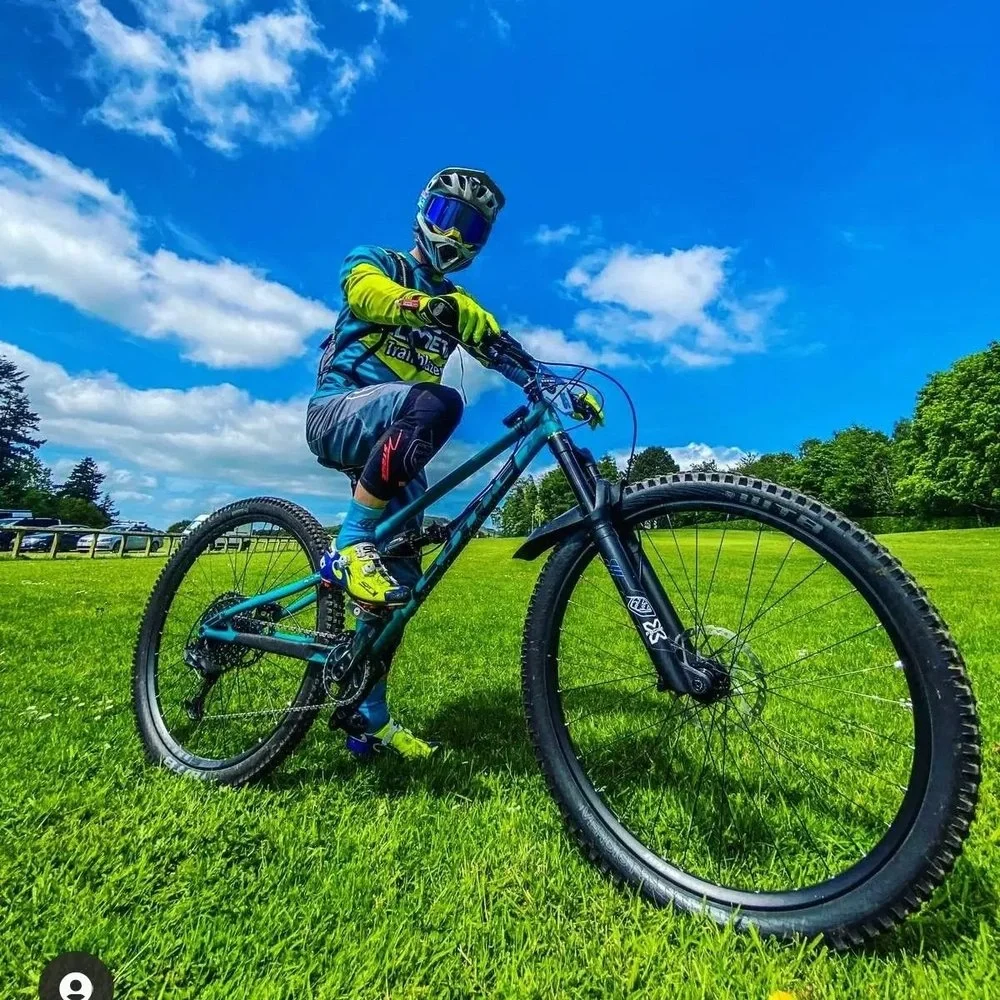 A person wearing a helmet, goggles, a yellow and blue cycling jersey, black shorts, and blue socks rides a mountain bike on a grassy field under a partly cloudy blue sky.