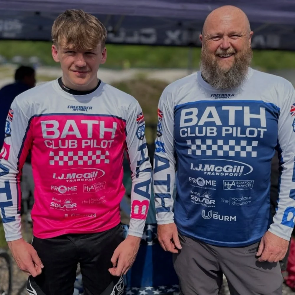 Two men standing side by side outdoors, wearing matching long-sleeve shirts with 'Bath Club Pilot' text and various sponsor logos. The man on the left is younger with short curly hair, wearing a pink shirt, and the man on the right is older with a ba