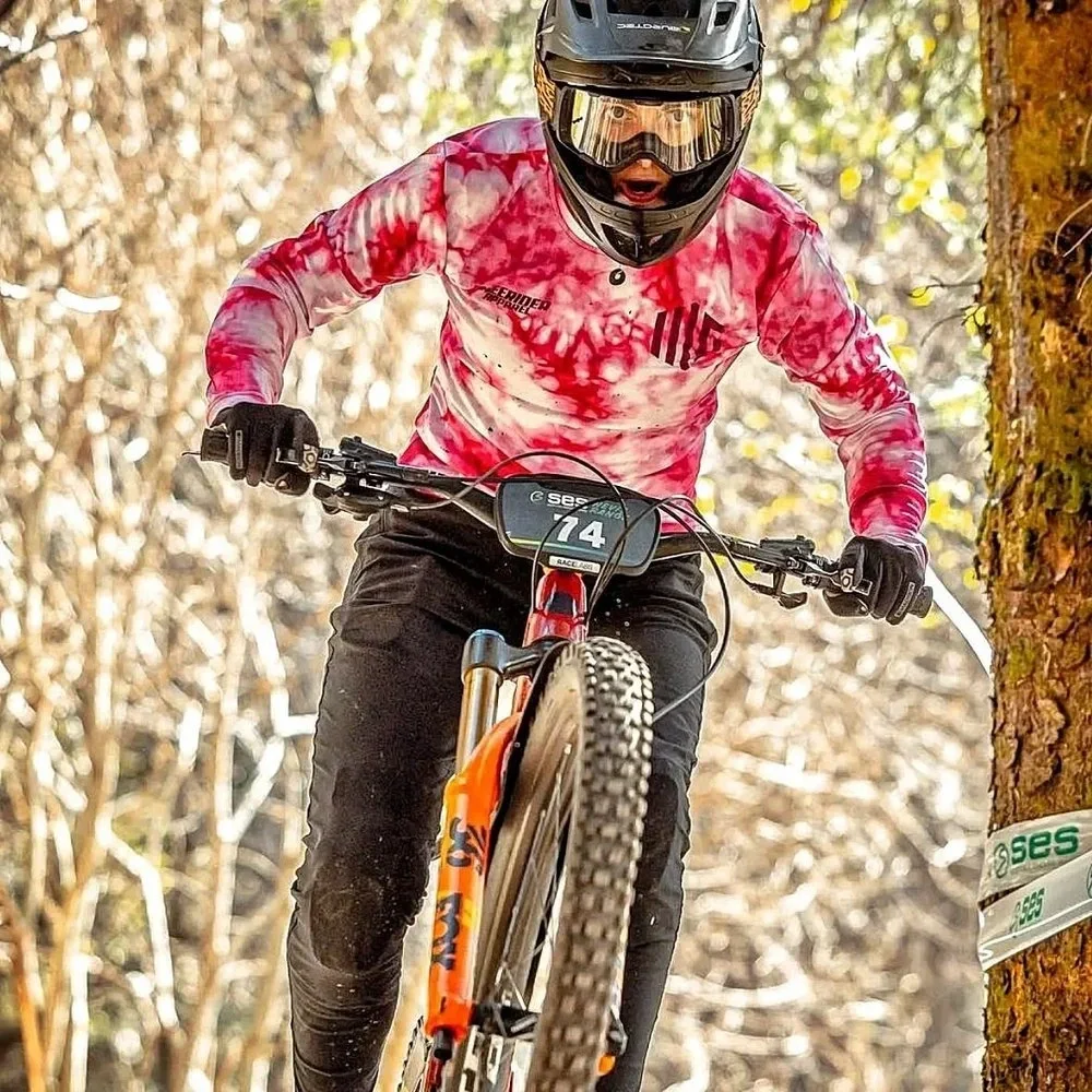 A mountain biker wearing a black helmet with goggles, a ladies cut  pink and white tie-dye long-sleeve jersey, black pants, and gloves, riding a mountain bike on a trail through a wooded area with autumn foliage.