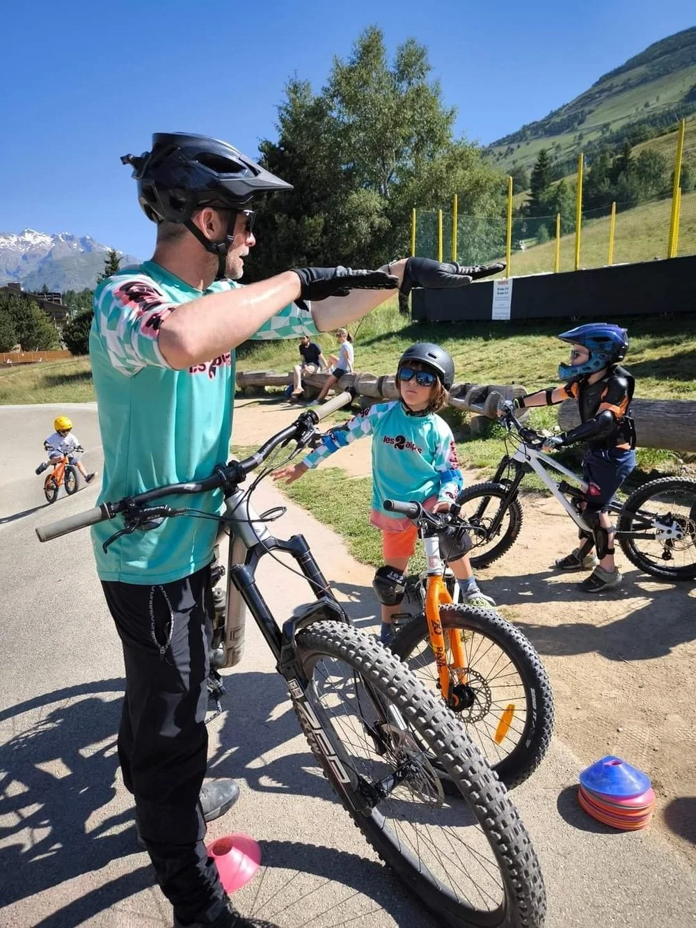 A man in a helmet and sunglasses giving a hand signal to two children on bicycles near a grassy area with mountains in the background on a sunny day.