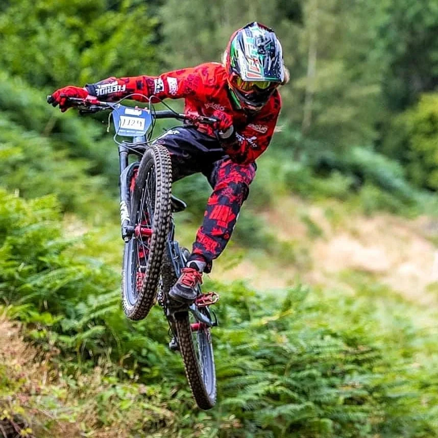 Mountain biker in red and black gear performing a jump on a dirt trail surrounded by green foliage.