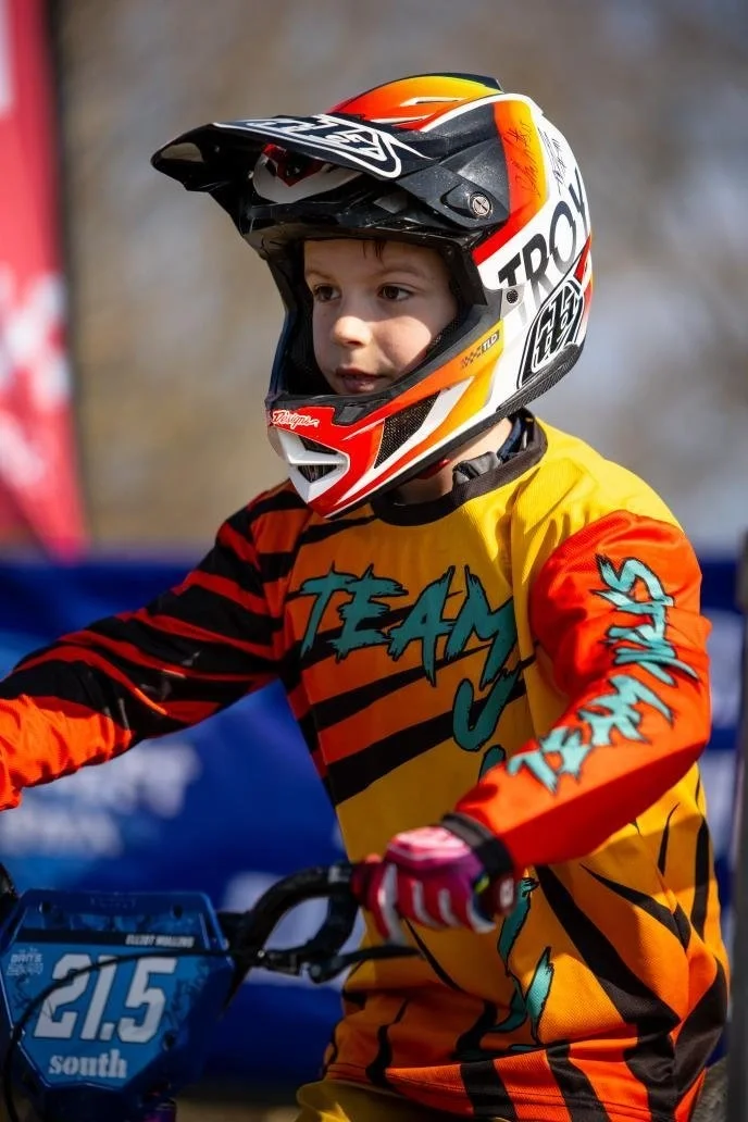 A young boy wearing a colorful motocross helmet and a yellow and orange racing jersey on a dirt bike, with the number 21.5 on the front.