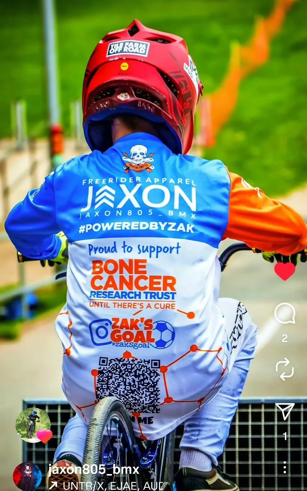 A BMX rider wearing a red helmet and a jersey with support logos for bone cancer research and Zak's goal, riding on an outdoor track with green grass and orange track barriers.