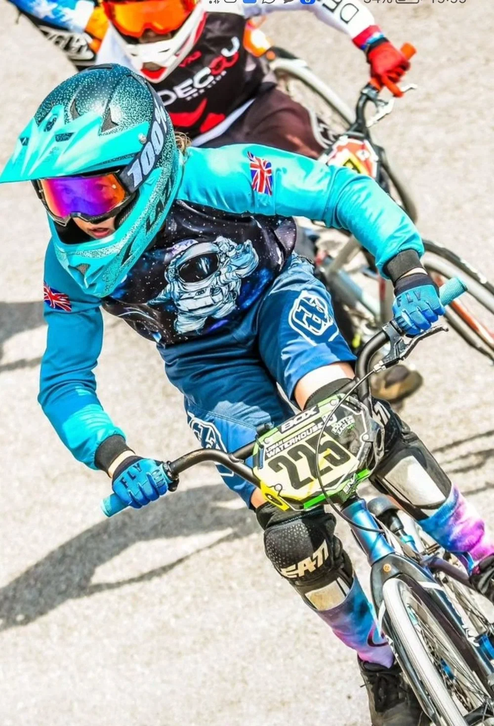 A young person riding a mountain bike wearing a blue helmet, tinted goggles, a space-themed jersey, blue gloves, and colorful leggings, participating in a cycling race.