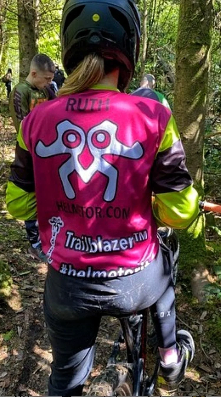 Person riding a mountain bike in the woods, wearing a pink and black helmet, and a pink, black, and neon yellow jacket with the name Ruth on the back.