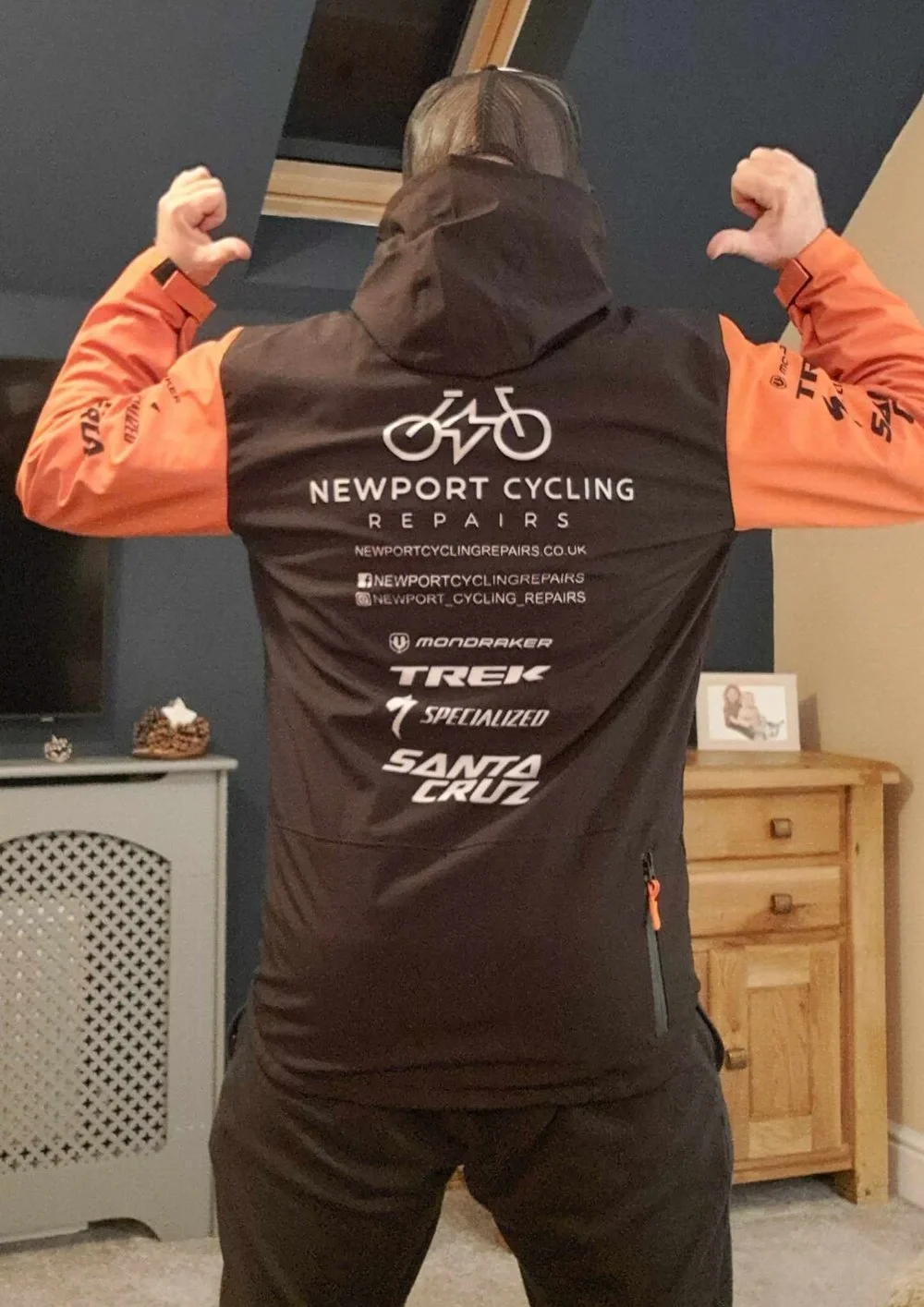 Person wearing a Newport Cycling Repairs jacket with logos and social media handles, flexing their muscles in a room with furniture.