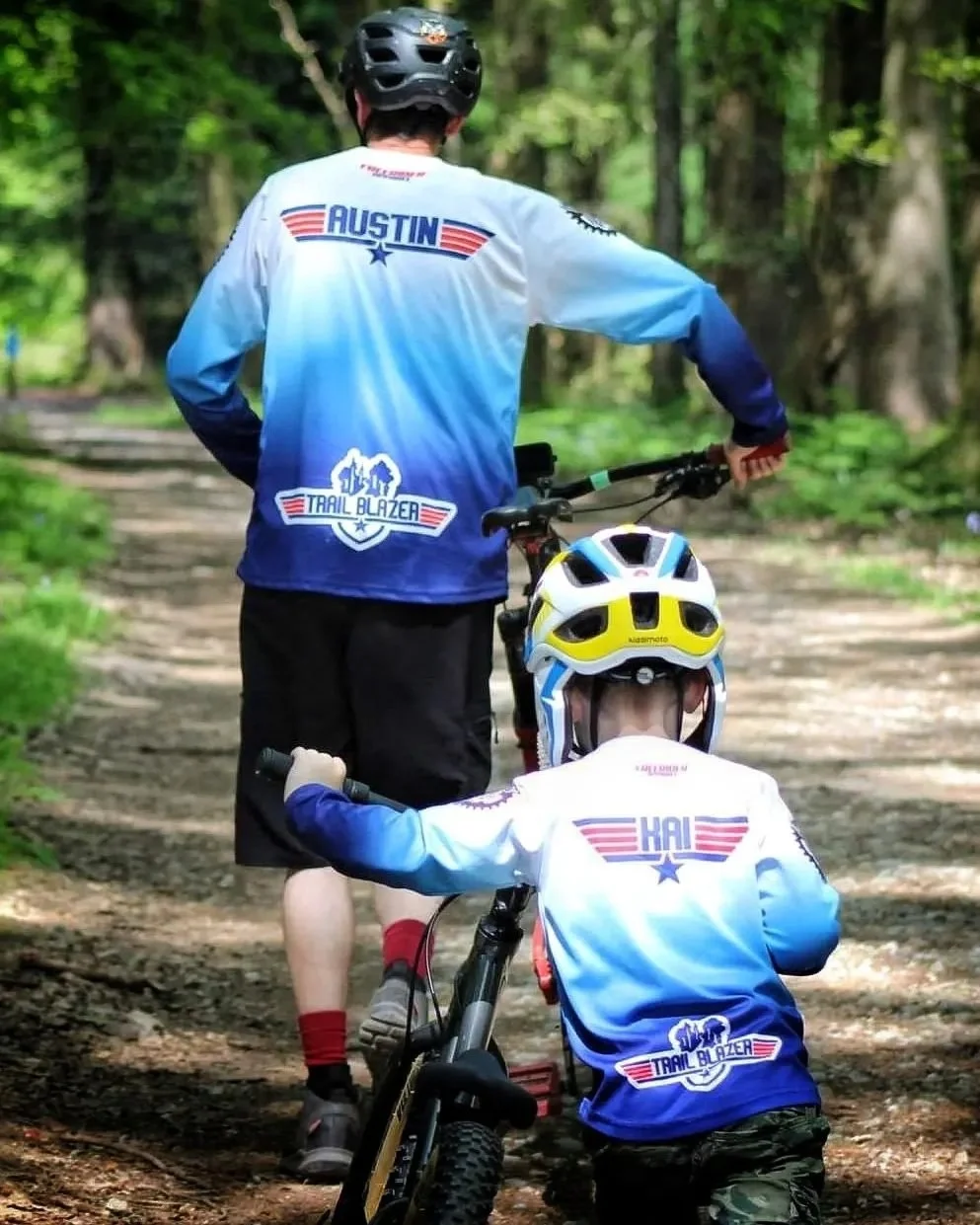 A man and a child in motocross gear riding mountain bikes on a forest trail.