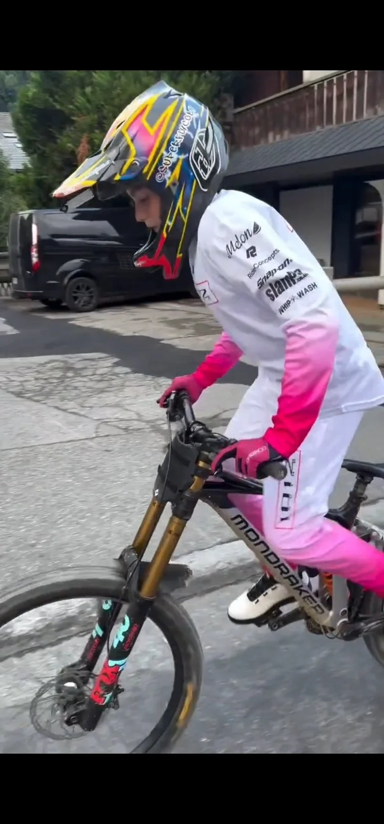 Motorcycle rider wearing a colorful helmet and a white and pink racing suit, riding a mountain bike on a paved street.