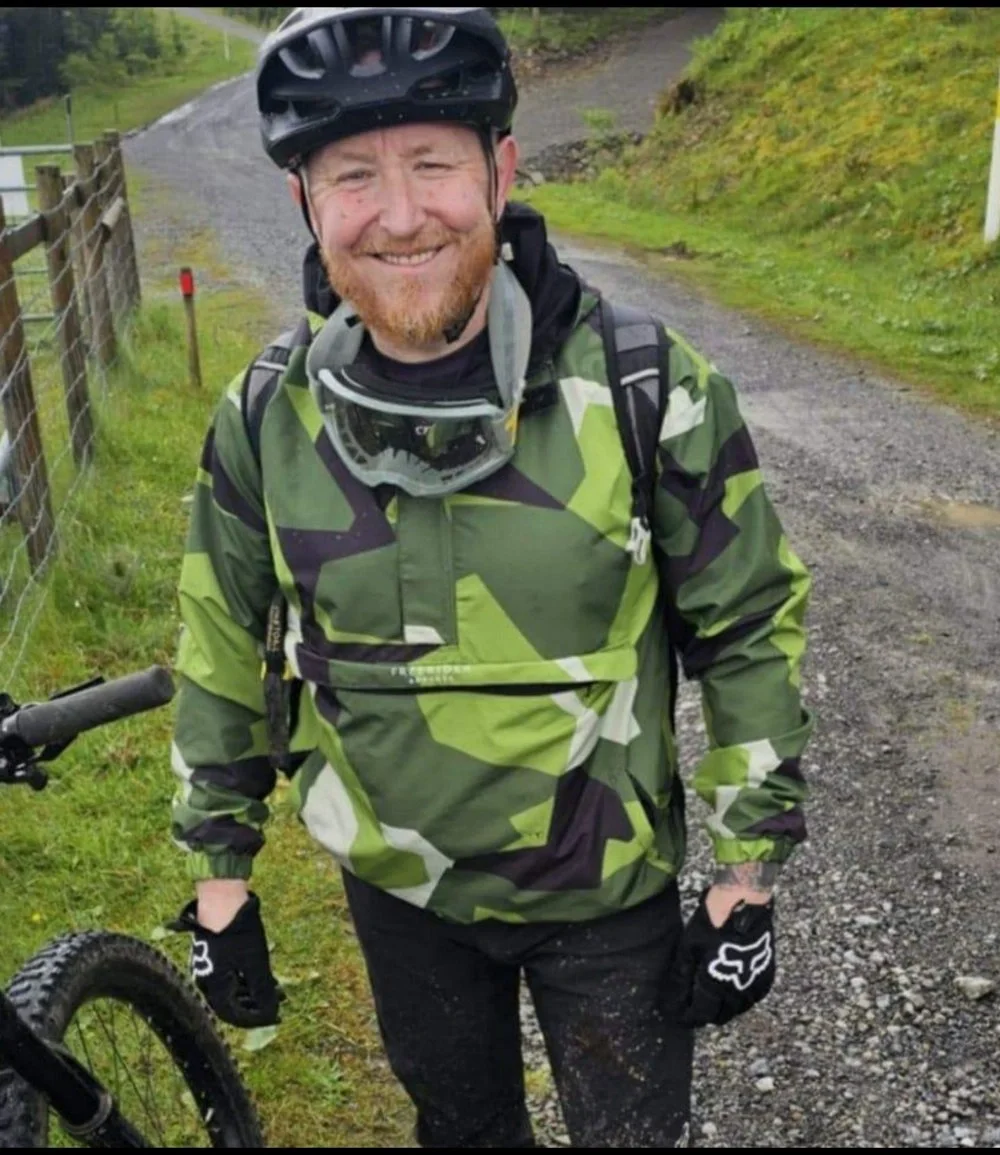 Freerider Apparel owner Shaun Evans wearing his custom camouflage waterproof mountain bike jacket and black gloves standing next to a mountain bike on a dirt trail.