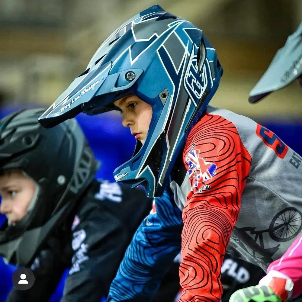 Young male motocross rider wearing blue and red helmet and gear, sitting on a dirt bike, focused on the race.