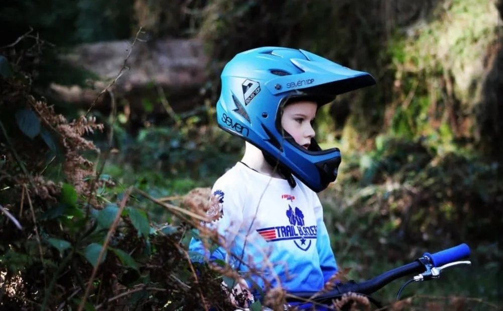 Young boy wearing a blue mountain biking helmet and a white long sleeve shirt with a bicycle graphic, standing in a forested area with bushes and trees.