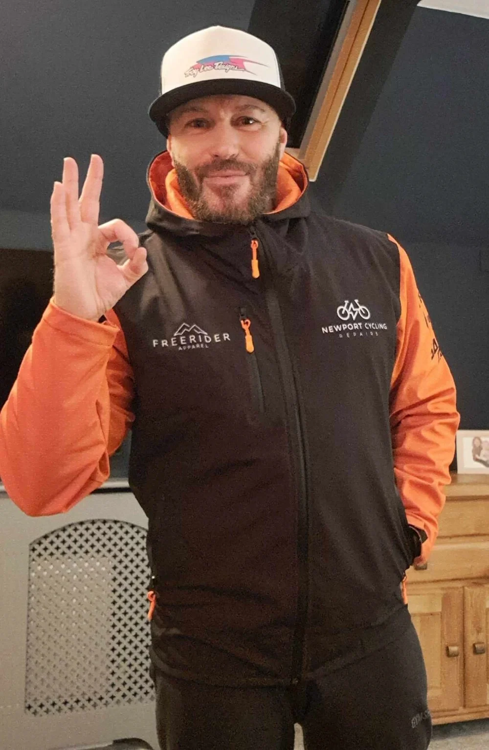 A man wearing a white cap with a logo, black and orange custom designed waterproof softshell MTB jacket with 'Freerider Apparel' and 'Newport Cycling' logos, making an OK sign with his right hand, smiling indoors.