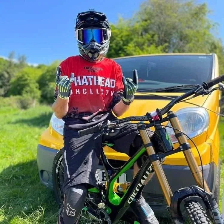 Child wearing full-face helmet, red shirt, and gloves standing with a mountain bike in front of a yellow car outdoors, with green trees and blue sky in the background.