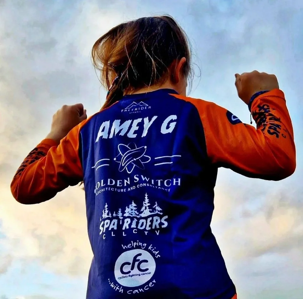 Back of young girl in orange and blue sports jersey with logo and text, flexing arms against cloudy sky background.