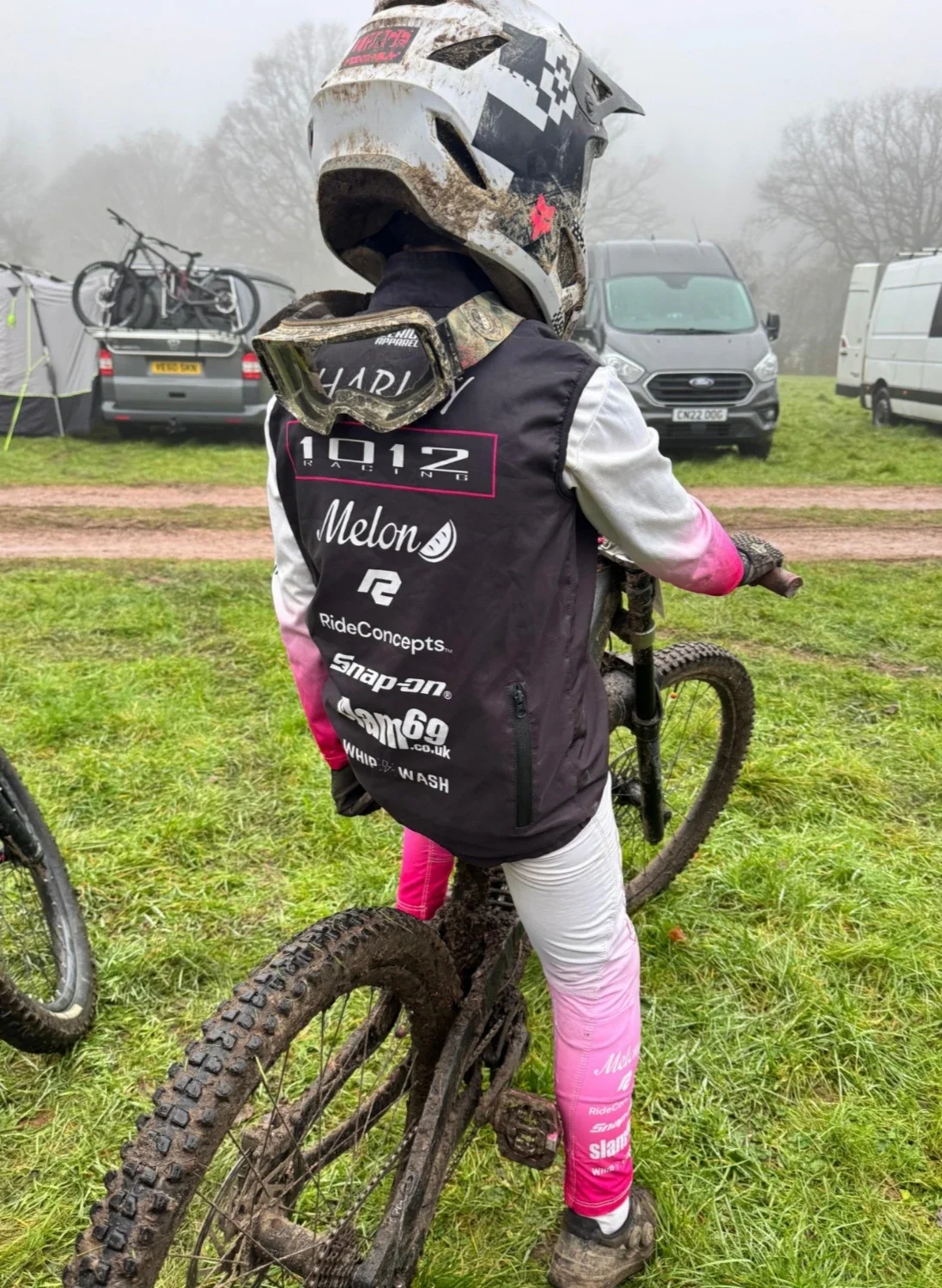 A person dressed in biking gear with a helmet and goggles, standing with a muddy mountain bike on a grassy field with tents and vehicles in the background.