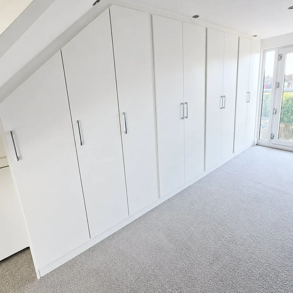 A fitted loft wardrobe installation in a crisp white finish, providing seamless storage across an angled bedroom wall.