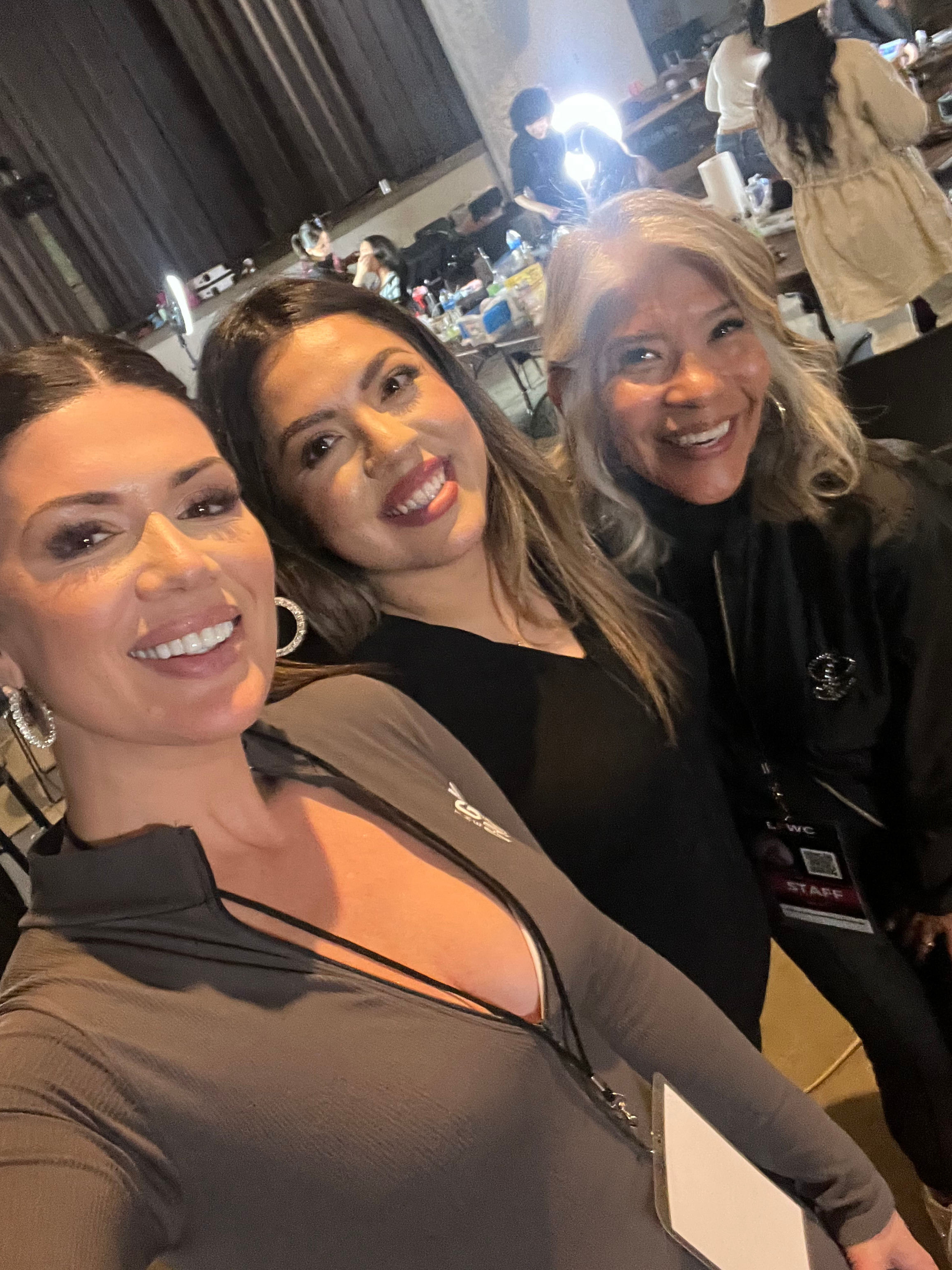 Three women taking a selfie in a busy kitchen or cafeteria. One woman in the foreground has dark hair and is smiling, the middle woman has light hair and is also smiling, and the woman on the right has light curly hair and is smiling broadly. In the background, other people are preparing food or working, with tables and kitchen supplies visible.