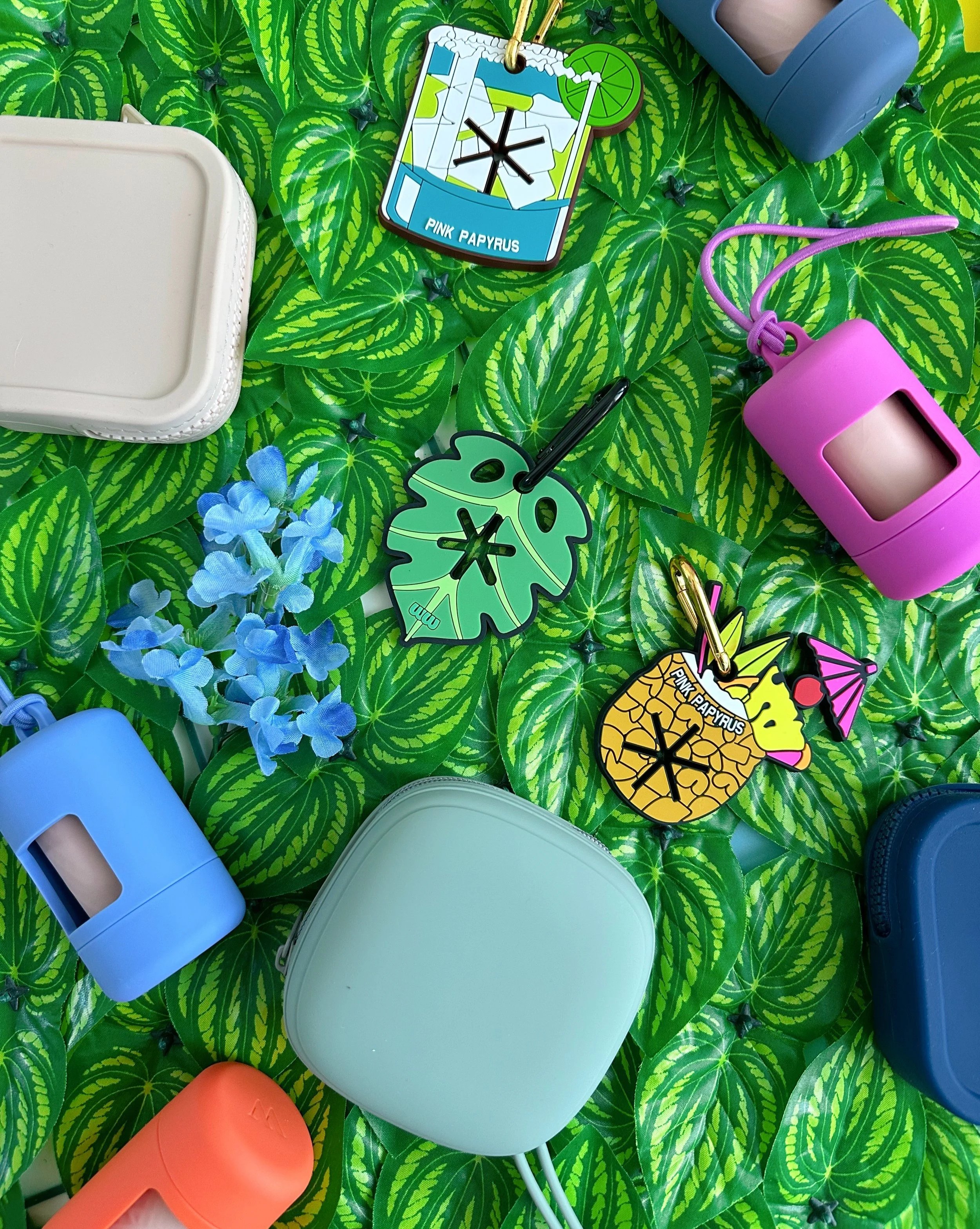 Collection of small, colorful safety and waterproof cases with clips, shaped like pineapples, monstera leaves, and other fun designs, arranged on a leafy green tablecloth. Some are open, revealing hand sanitizer bottles inside.