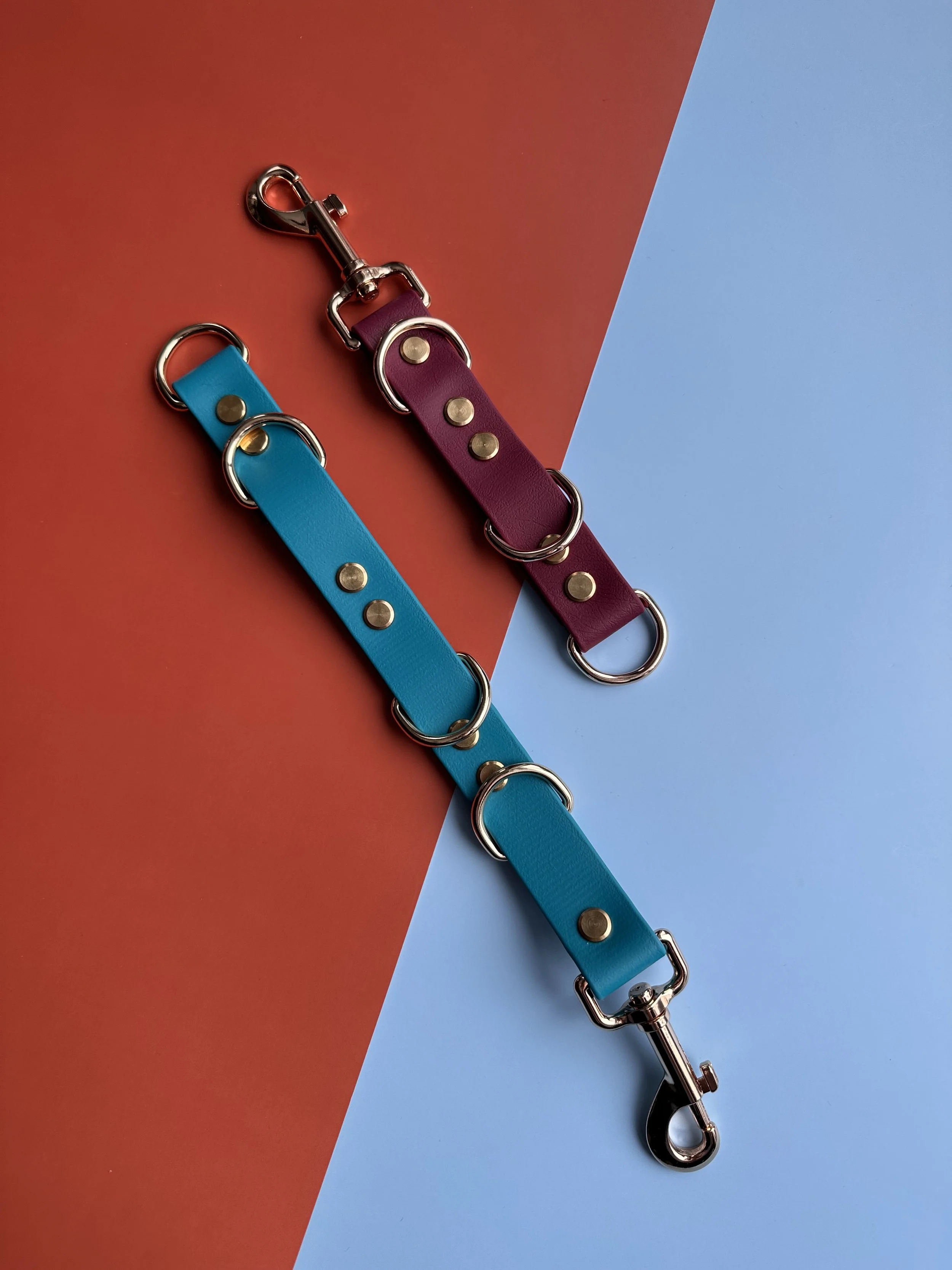 Two colorful leather dog leashes, one blue and one purple, with metal hardware on a split background of orange and blue.