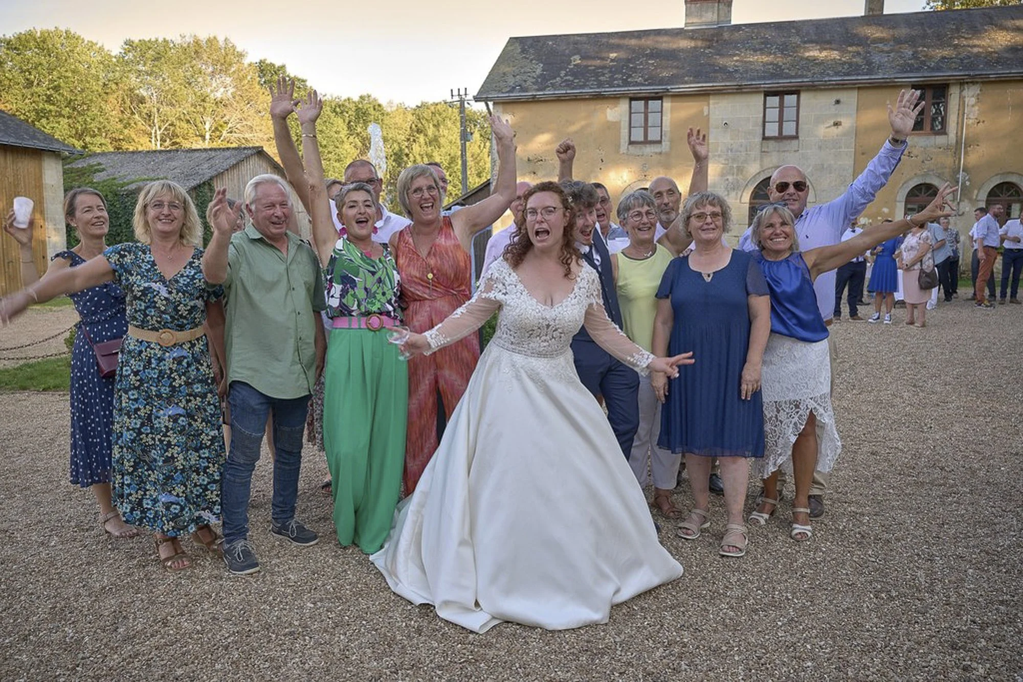 Groupe de personnes souriantes et levant les mains lors d'une célébration en plein air devant une vieille ferme en pierre, une femme en robe de mariage au centre.