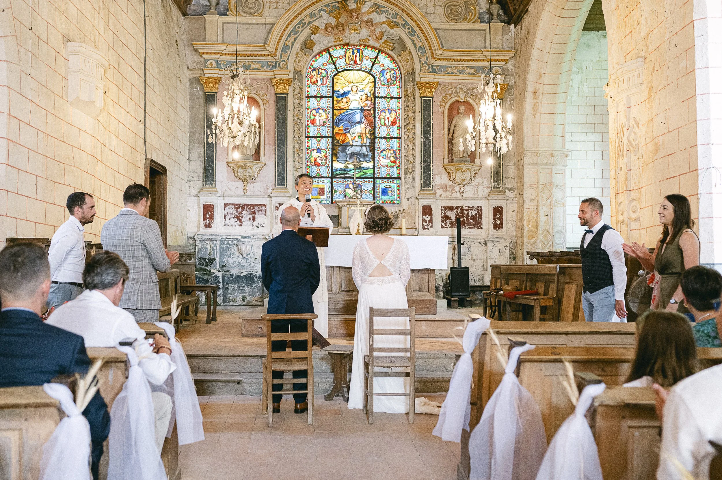 Cérémonie de mariage dans une église avec un couple devant le prêtre, invités assis et décorations classiques derrière un grand vitrail coloré représentant Jésus avec des anges.