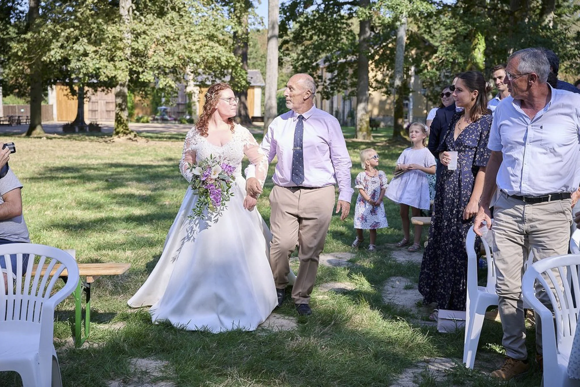 Un groupe de personnes lors d'un mariage en plein air, avec des arbres en arrière-plan. La mariée, en robe blanche, tient un bouquet de fleurs et marche avec un homme âgé. D'autres invités, quelques enfants et adultes, regardent et participent à la c