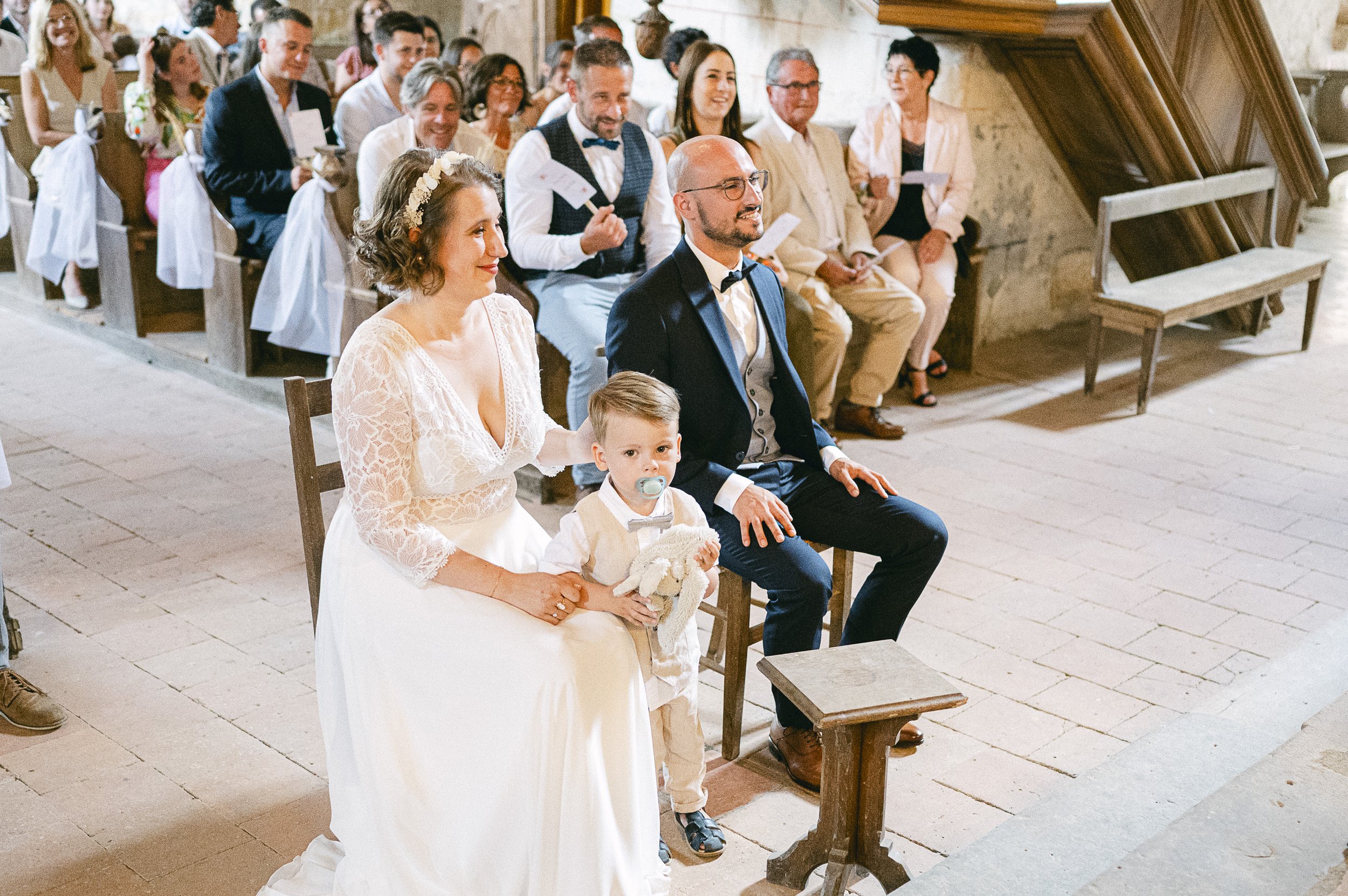 Cérémonie de mariage dans une église avec le couple marié assis devant, entouré de invités assis dans les bancs en bois.