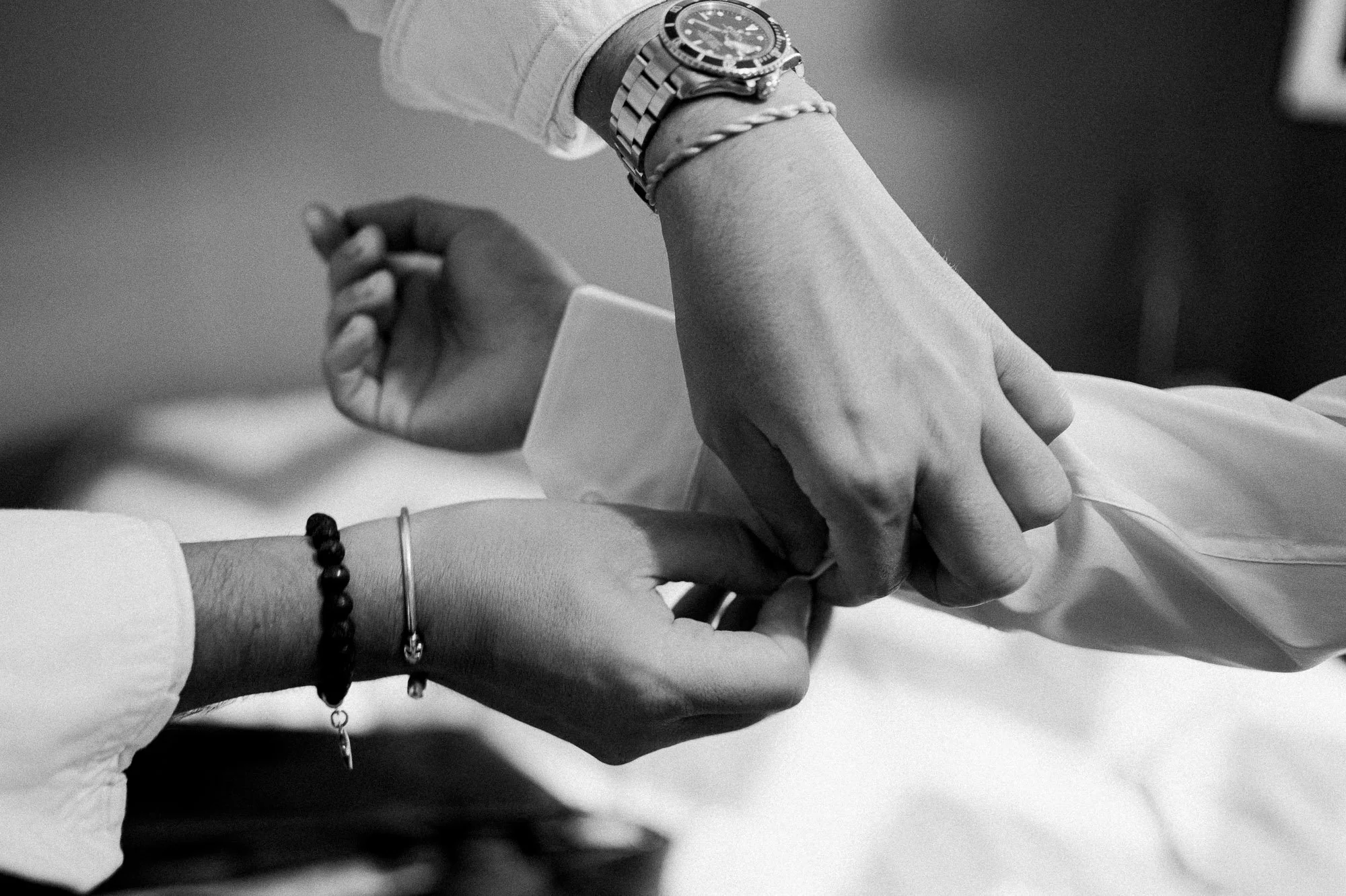Une personne porte une montre-bracelet en métal et une chaîne, et tient la main d'une autre personne avec un bracelet à perles, dans une ambiance en noir et blanc.