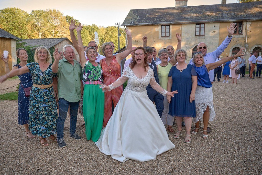 Groupe de personnes en fête, souriantes et levant les bras, avec une femme en robe de mariée au centre.