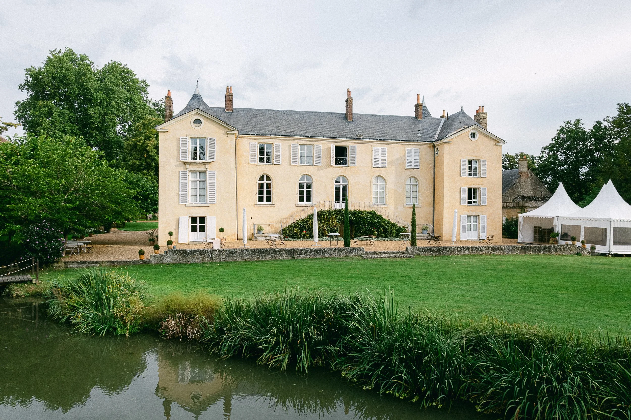 Un grand château de style classique avec des murs beige clair et des fenêtres à volets blancs, entouré d’un parc verdoyant avec un étang au premier plan.